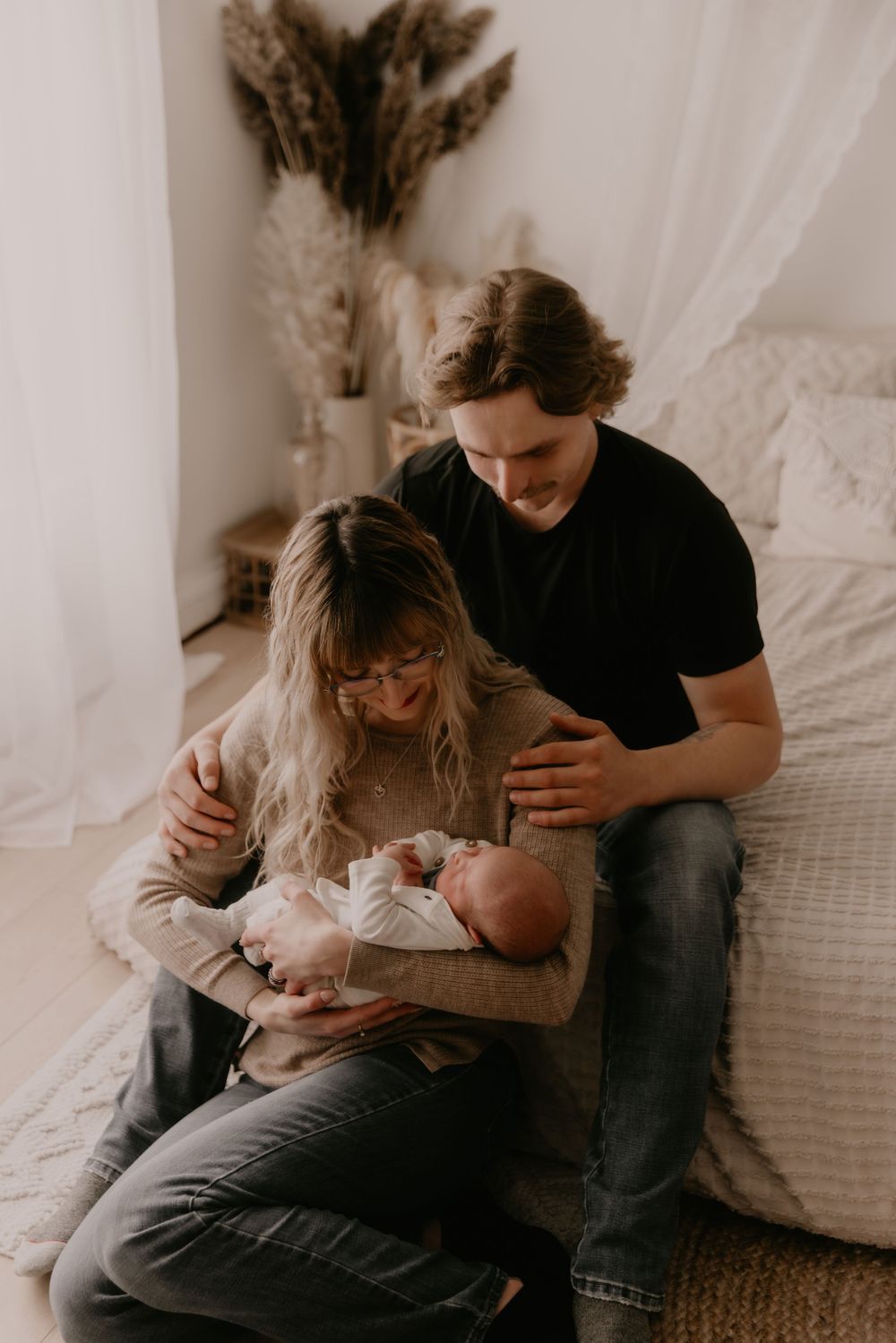 Family cuddles with newborn baby in a cozy bedroom with soft white curtains and natural lighting.