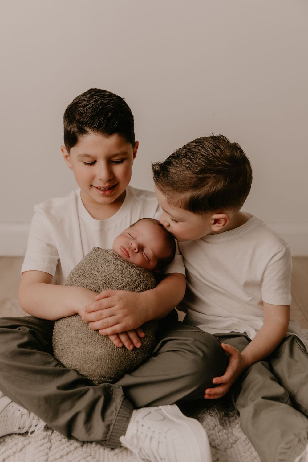 Two young siblings in white shirts and gray pants share tender moments while sitting together on a light colored surface.