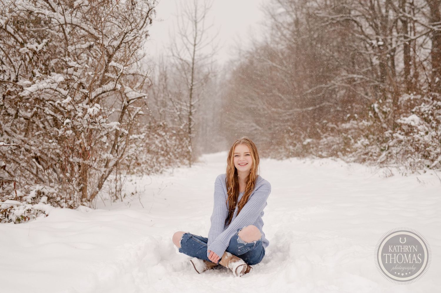 A person sits cross-legged in snow wearing a gray sweater and jeans on a wintry forest path.