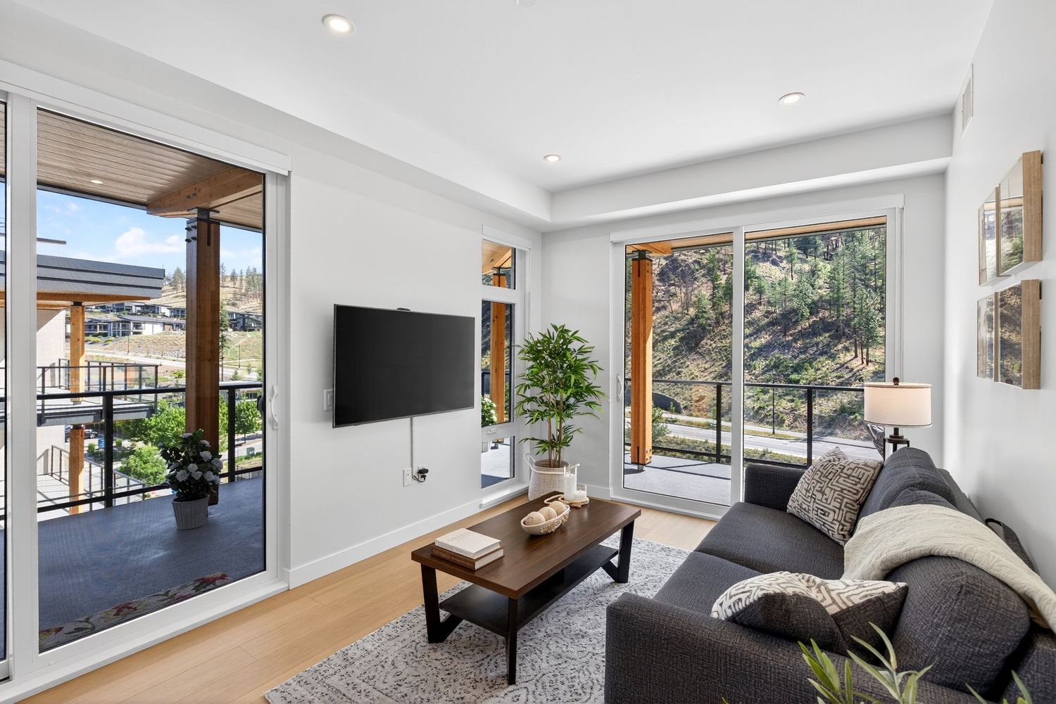 Modern living room with gray sofa, wall-mounted TV, wood coffee table, and large windows overlooking lush greenery.