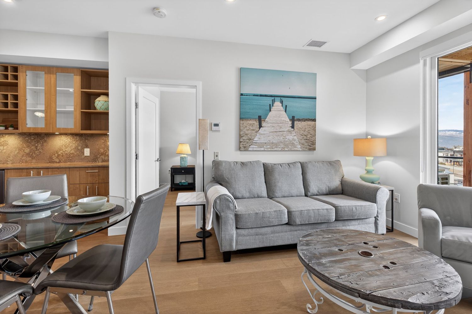 Modern living room with gray sofa, glass dining table, wooden coffee table, and ocean artwork in a bright apartment.
