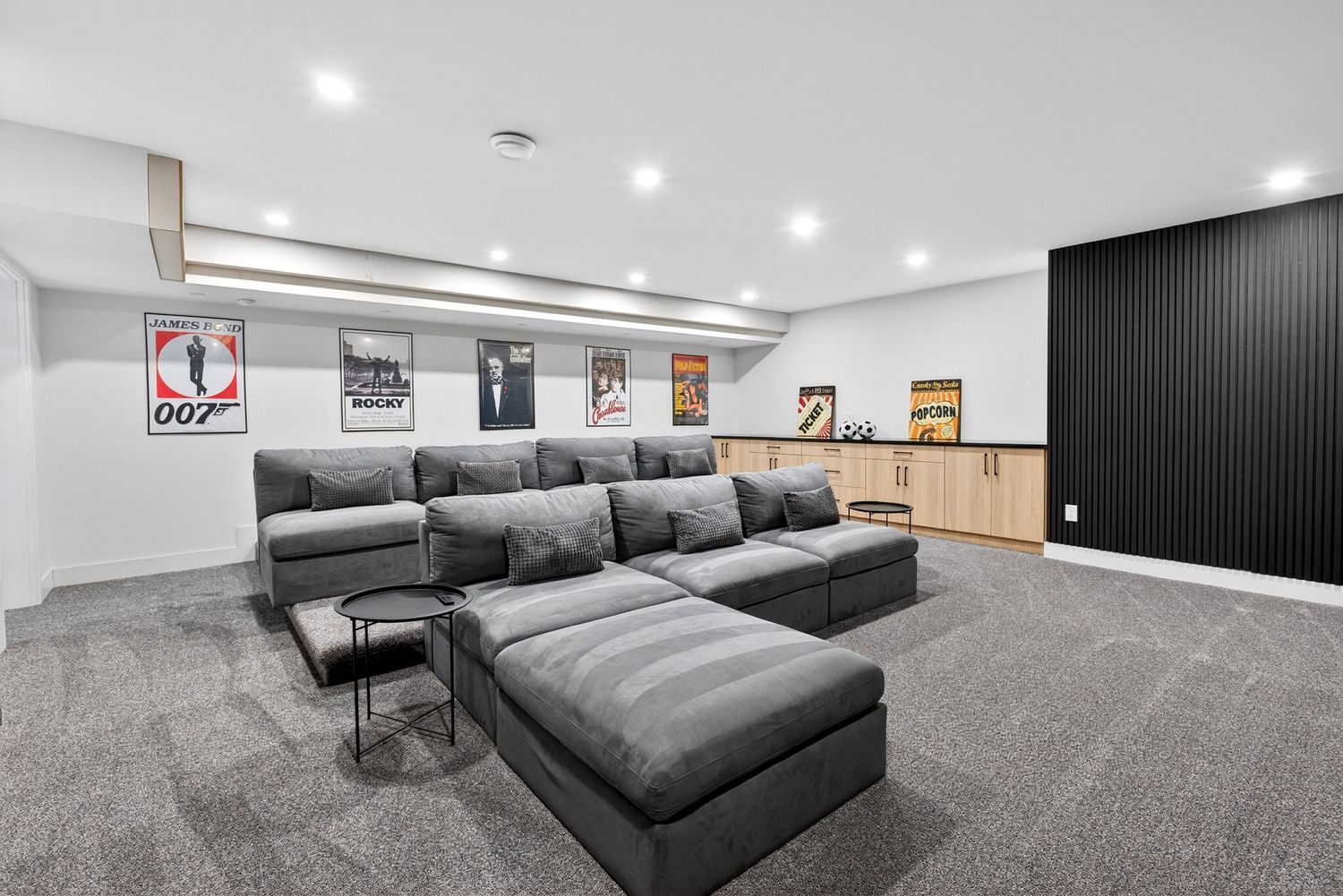 Modern basement home theater with gray sectional sofa, James Bond movie posters, black accent wall, and recessed lighting.