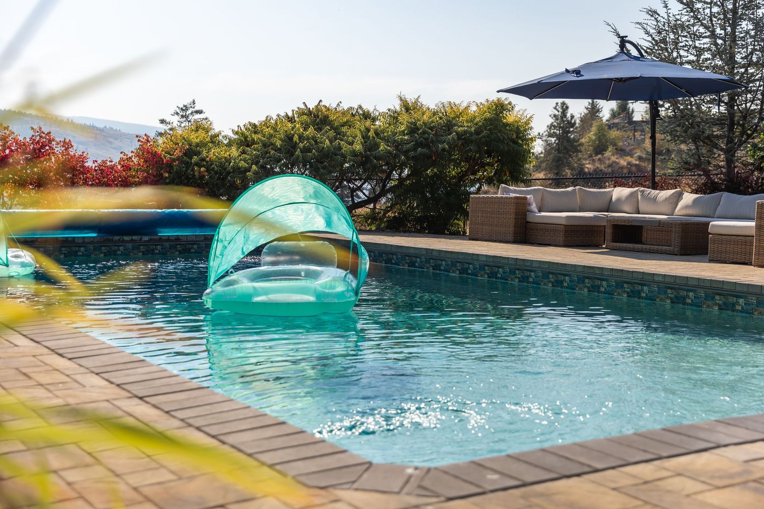 Modern swimming pool with blue water and stone patio surrounded by lush landscaping and lounge at a Kelowna Aribnb