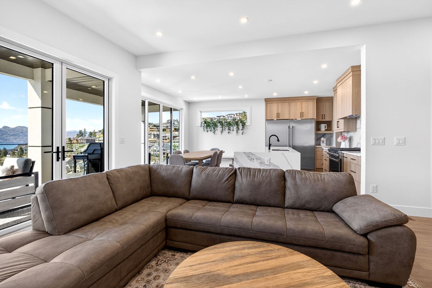 Modern open-concept living room with brown leather sectional sofa, hardwood floors, and mountain views through large windows.