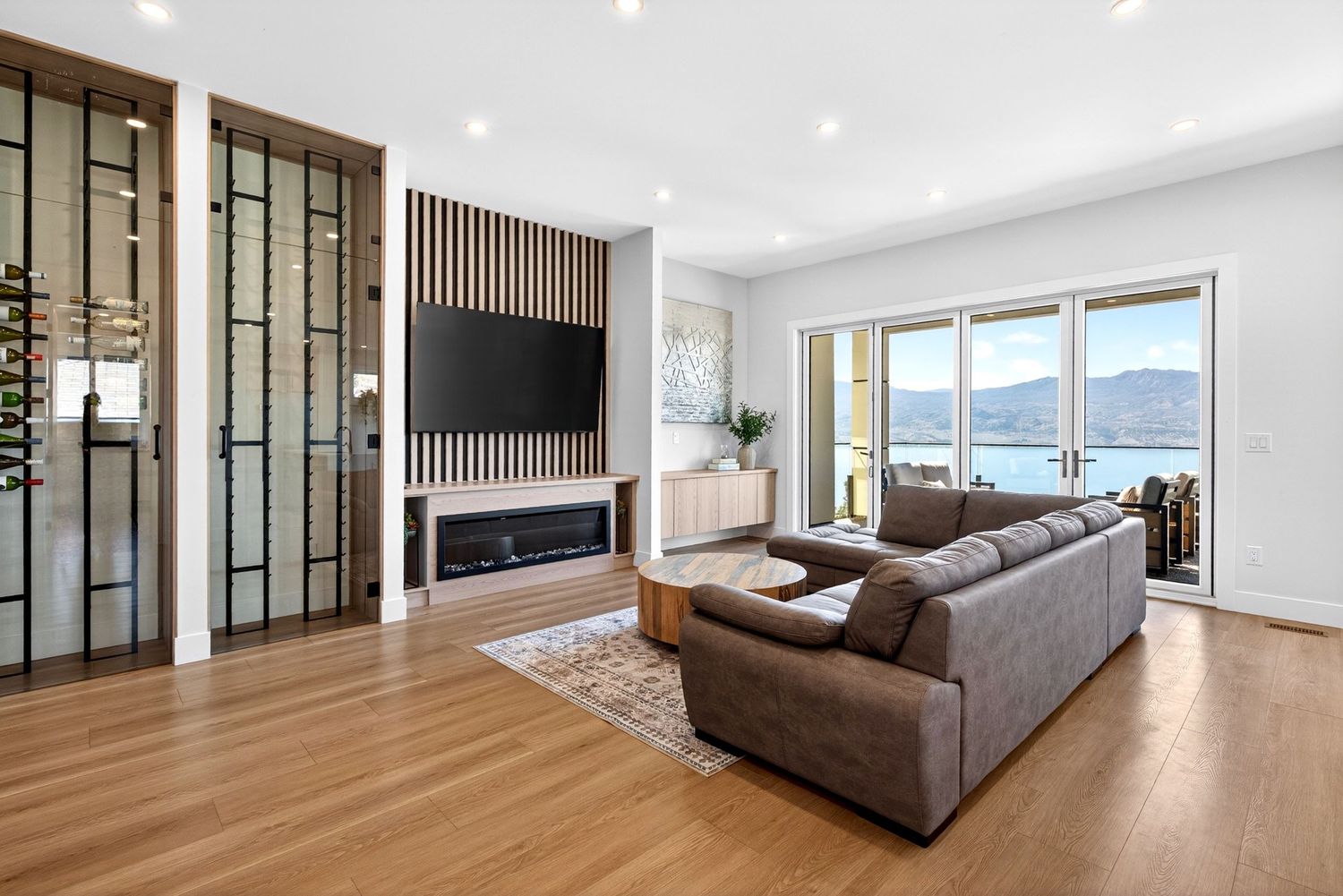 Modern living room with hardwood floors, leather sofa, wall-mounted TV, fireplace, and stunning lake view through large windows.