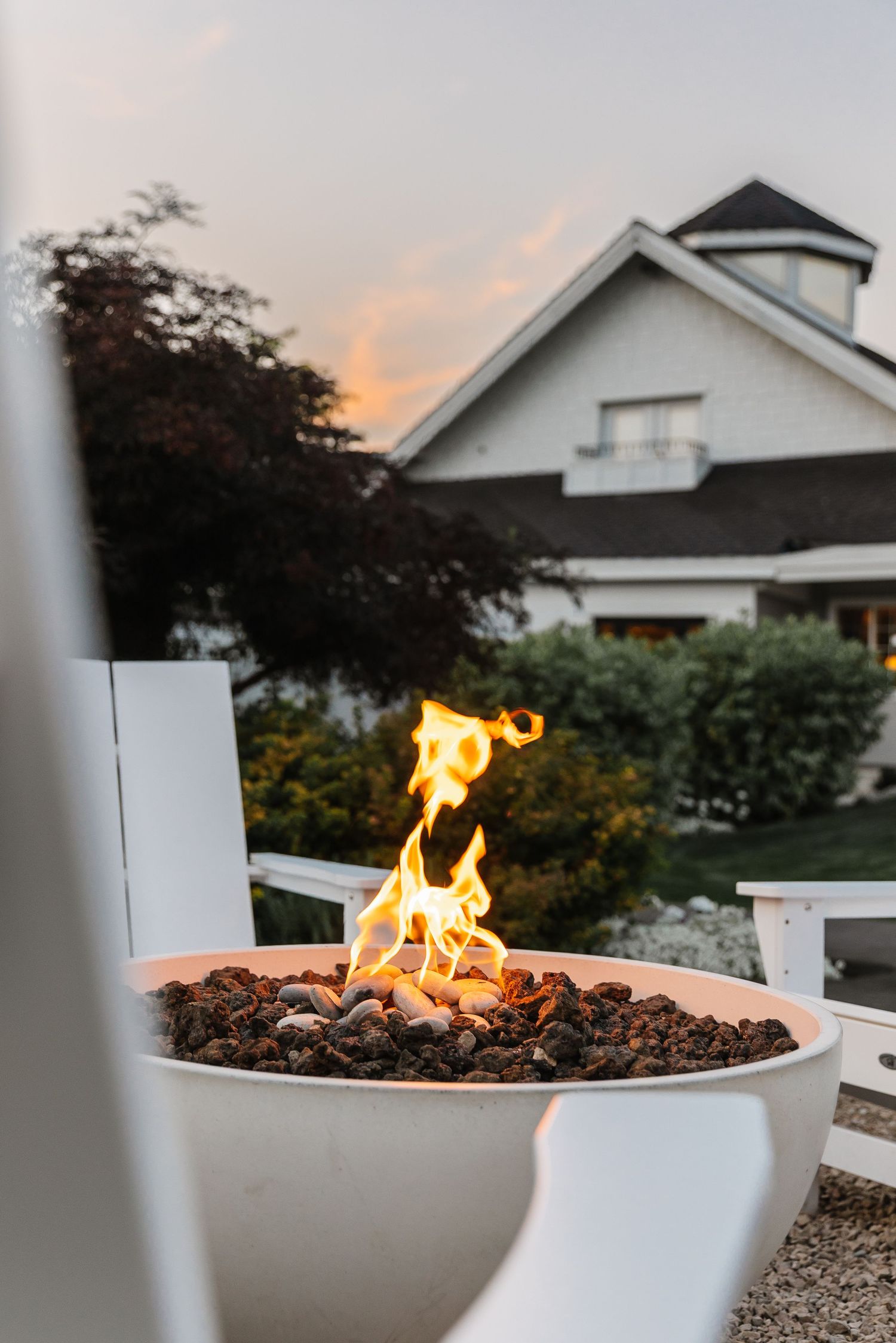 A cozy outdoor fire pit with dancing flames against a twilight sky and white house in the background.