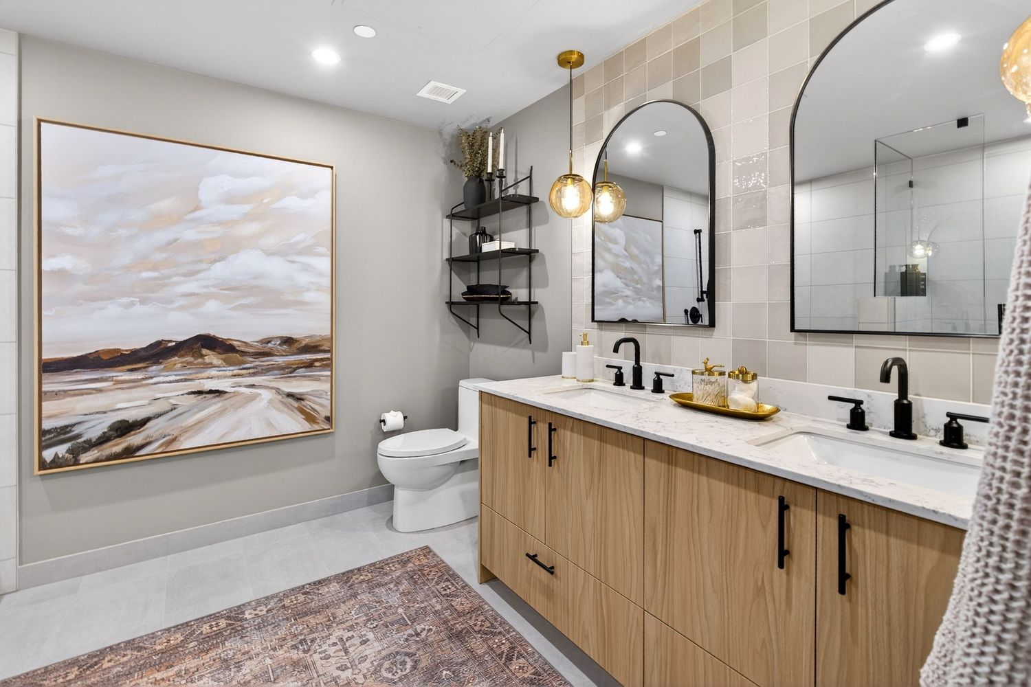 Modern bathroom with wood vanity, dual sinks, black fixtures, arched mirrors, pendant lights, and a landscape painting on gray walls.
