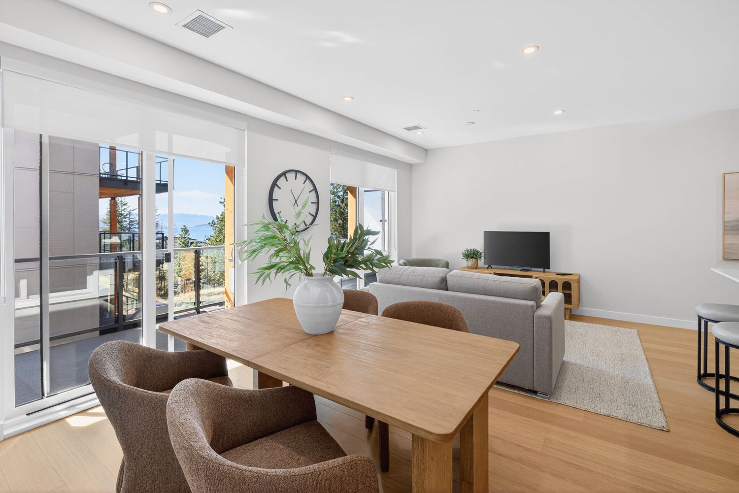 Modern open-plan dining and living room with wooden table, gray sofa, hardwood floors, and sliding doors to an ocean-view balcony.