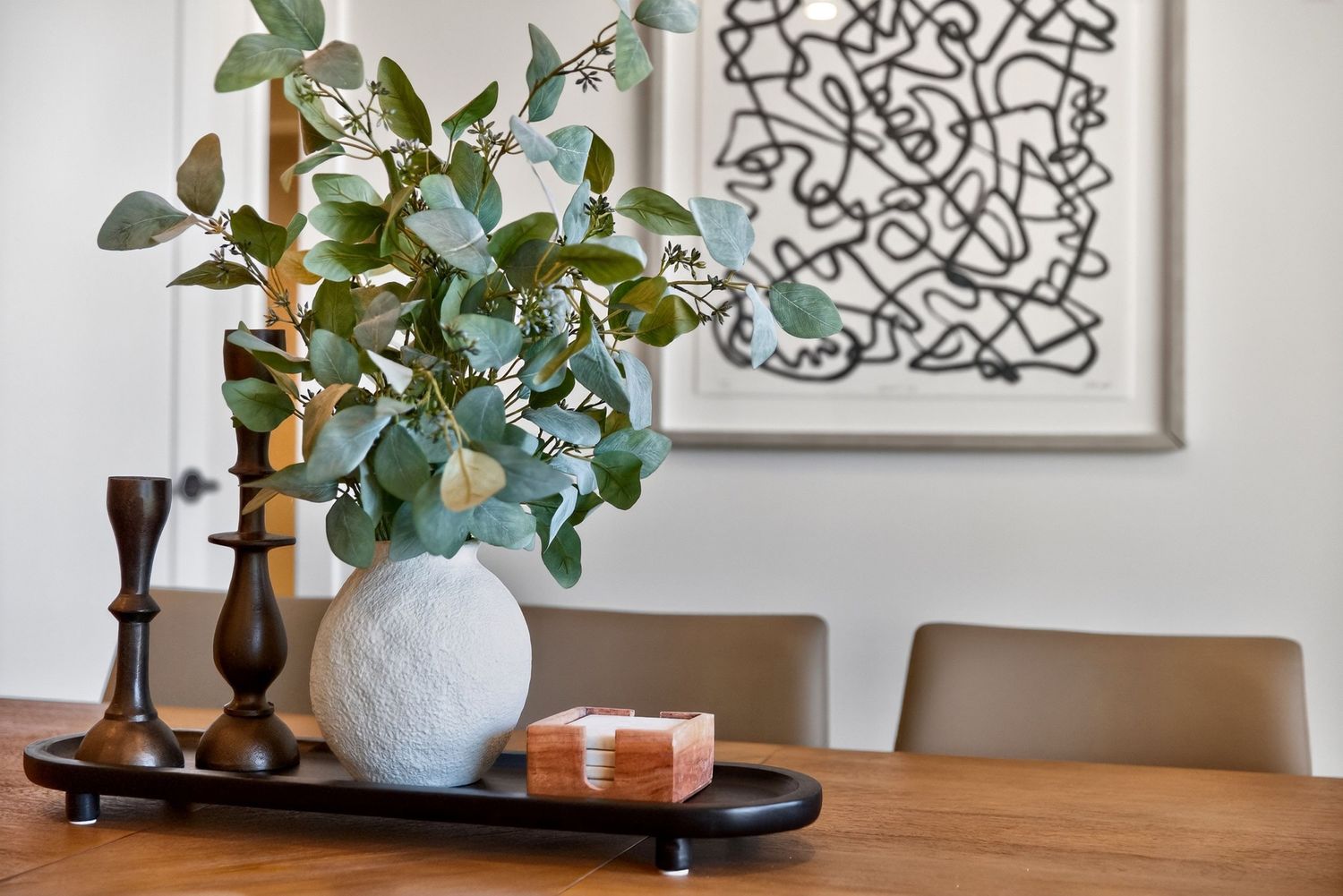 White textured vase with eucalyptus branches on a dark tray beside wooden candlesticks and abstract wall art.
