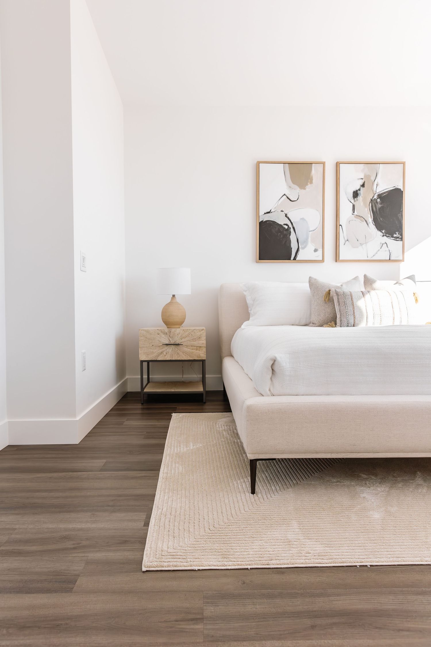 Minimalist bedroom with abstract art, beige bed frame, and modern nightstand on wood flooring.