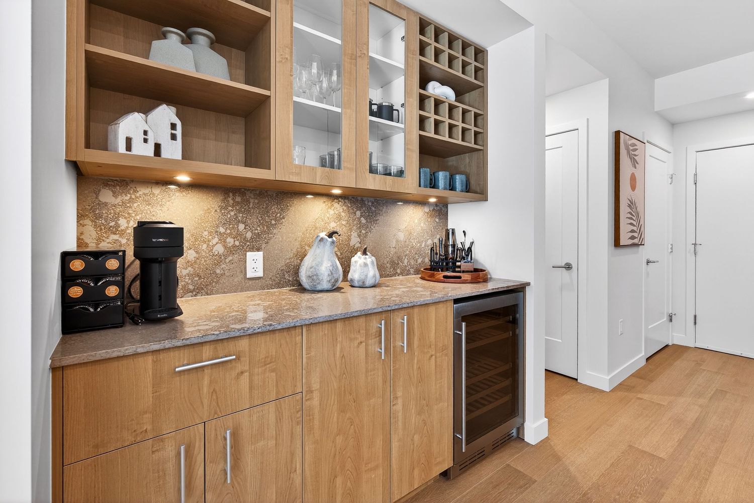 Modern kitchen coffee bar with wood cabinets, glass doors, and built-in appliances along a beige backsplash wall.