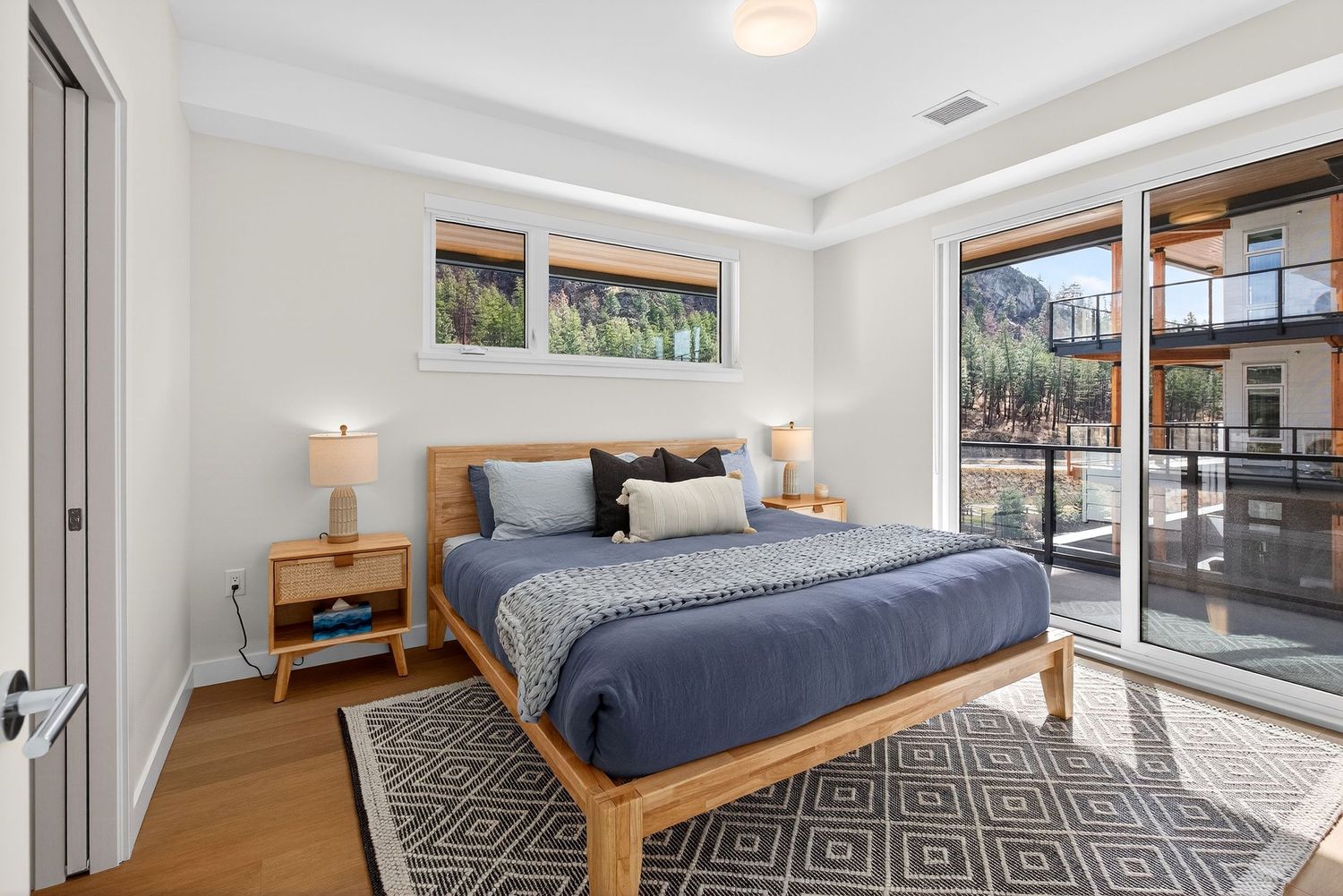 Modern bedroom with wooden bed frame, blue bedding, geometric rug, and large windows offering natural light.