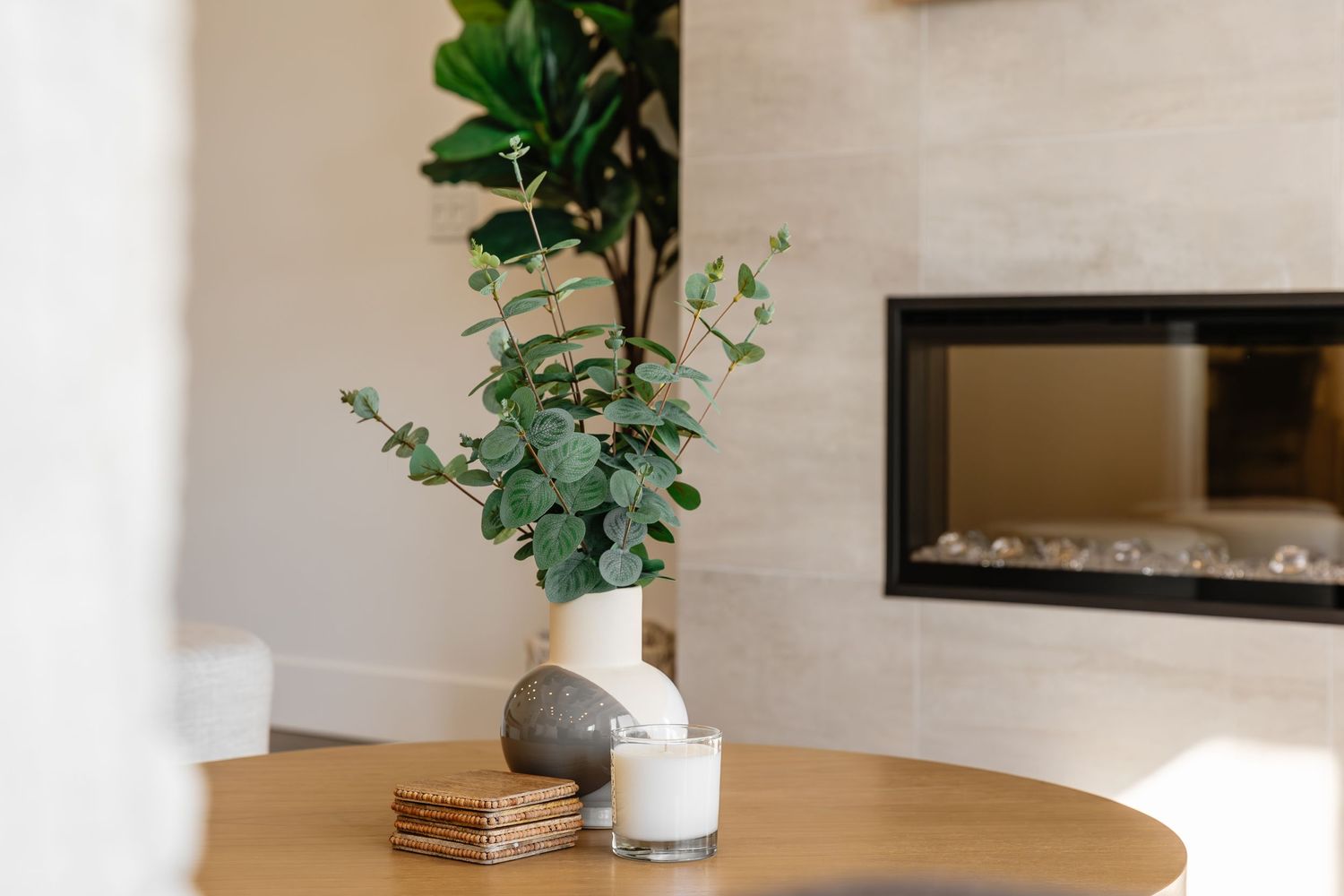 Decorative vase with eucalyptus stems on wooden table near modern fireplace.