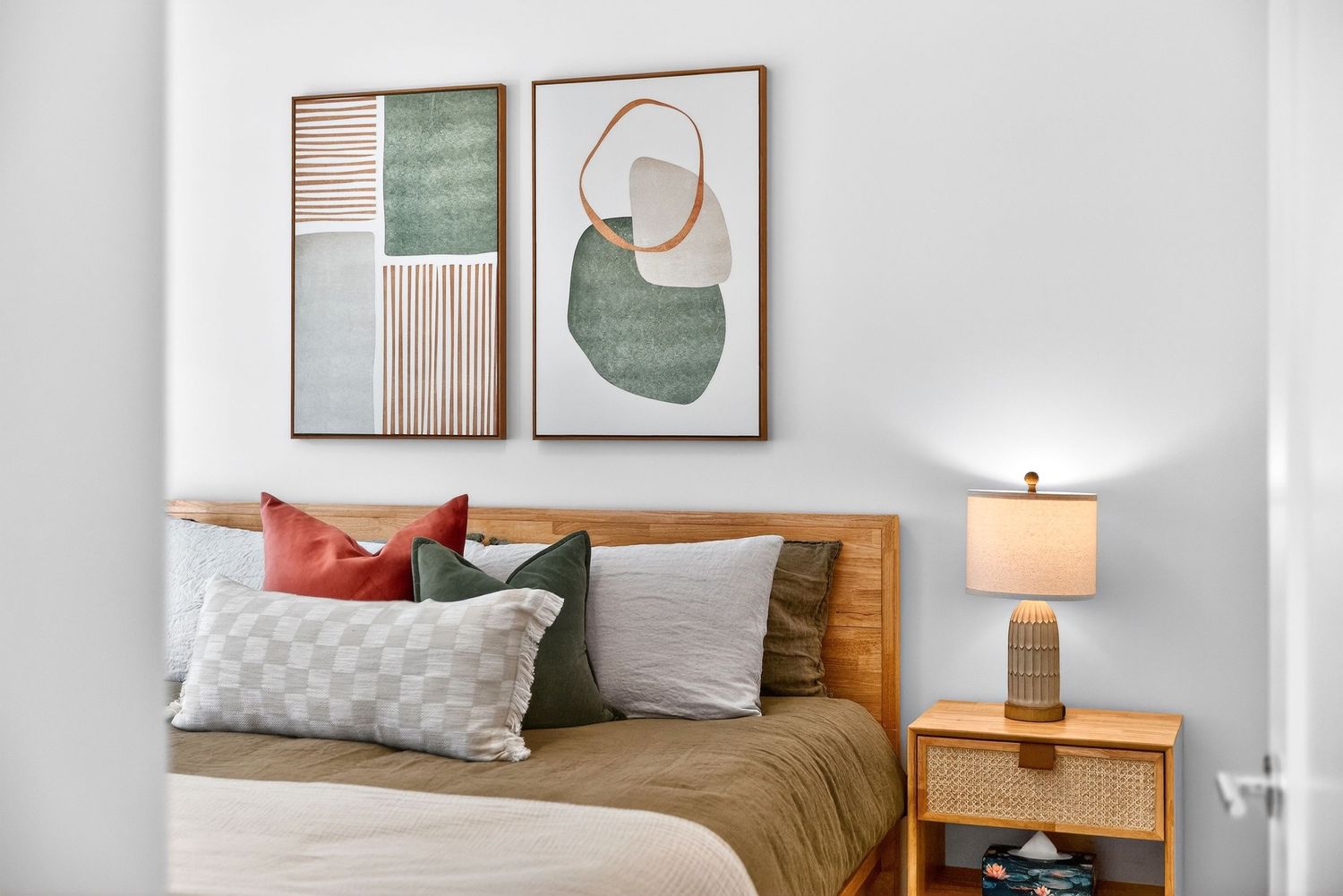 Modern bedroom with wooden bed frame, abstract wall art, and earth-toned bedding against a white wall.