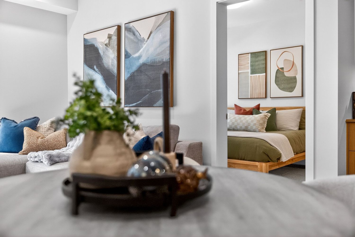 Modern living room with blue accent pillows and abstract art, leading to a cozy bedroom with green bedding.