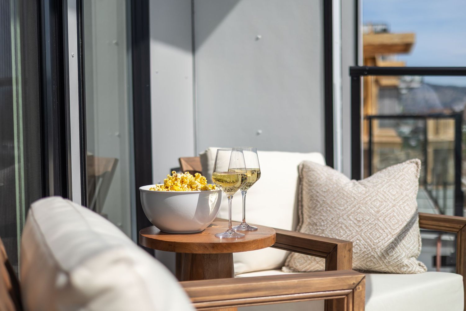 Cozy balcony seating area with two white wine glasses, a bowl of snacks on a wooden side table, and plush cushioned chairs.