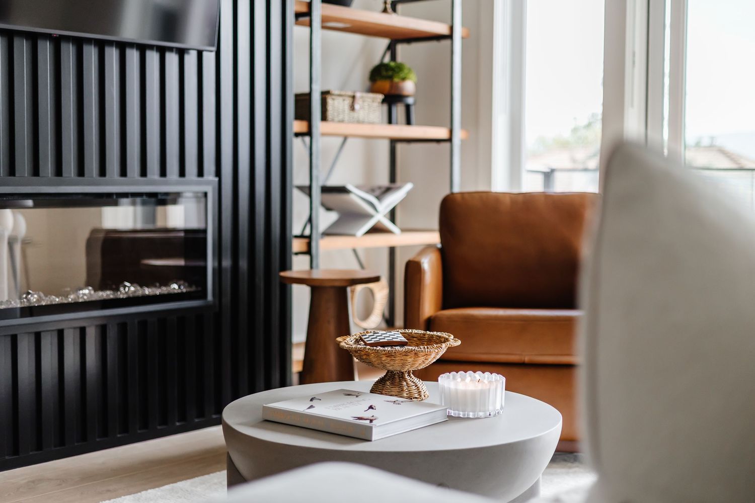 Modern living room with white round coffee table, gold bowl, candle, black fireplace, and brown leather armchair near window.