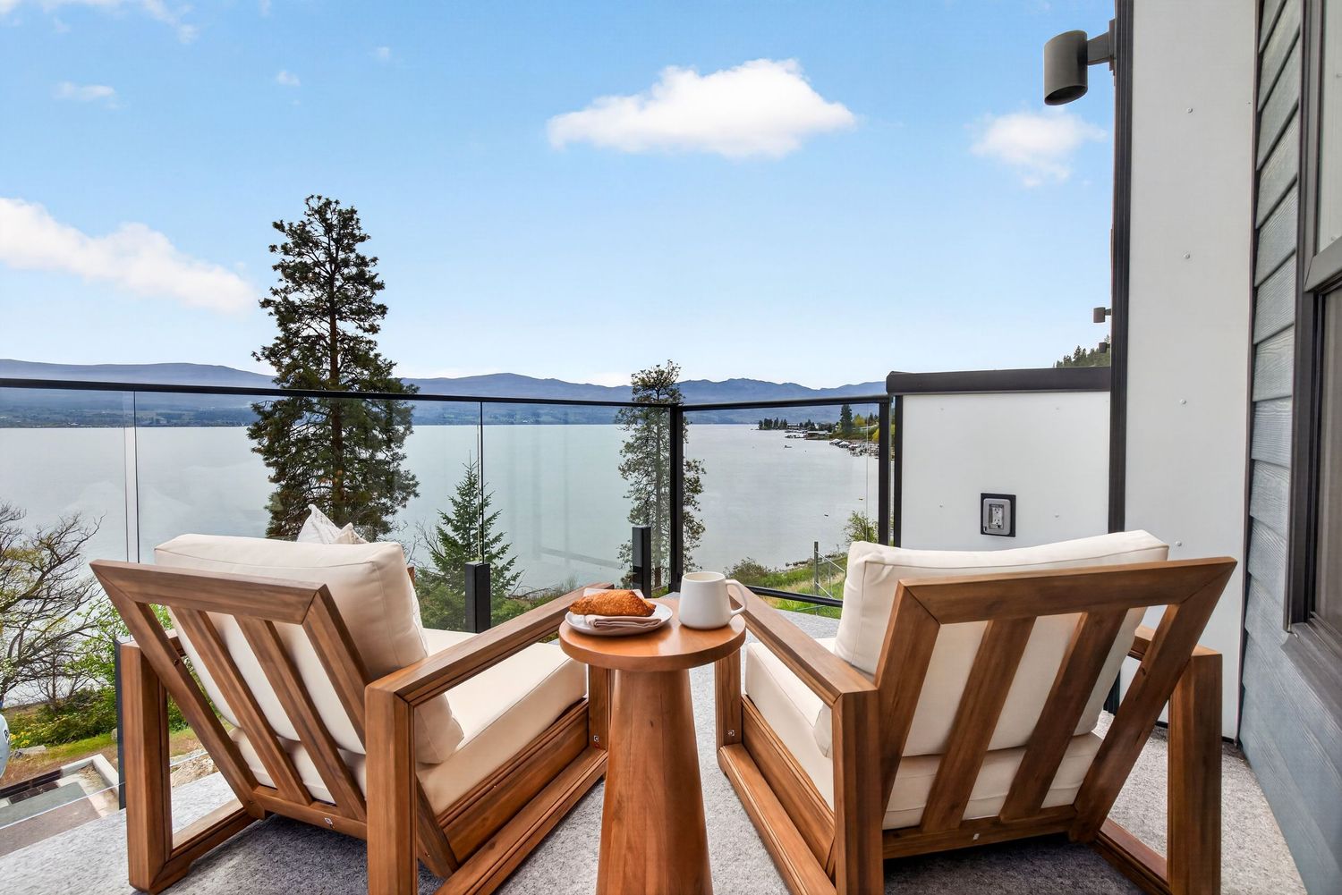 Luxury balcony with teak chairs and glass railing overlooking a serene lake and evergreen trees under a blue sky.