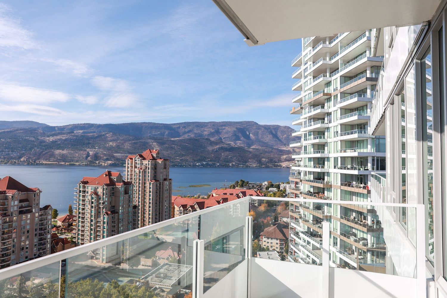 Glass balcony overlooking city skyline with mountains and high-rise buildings.