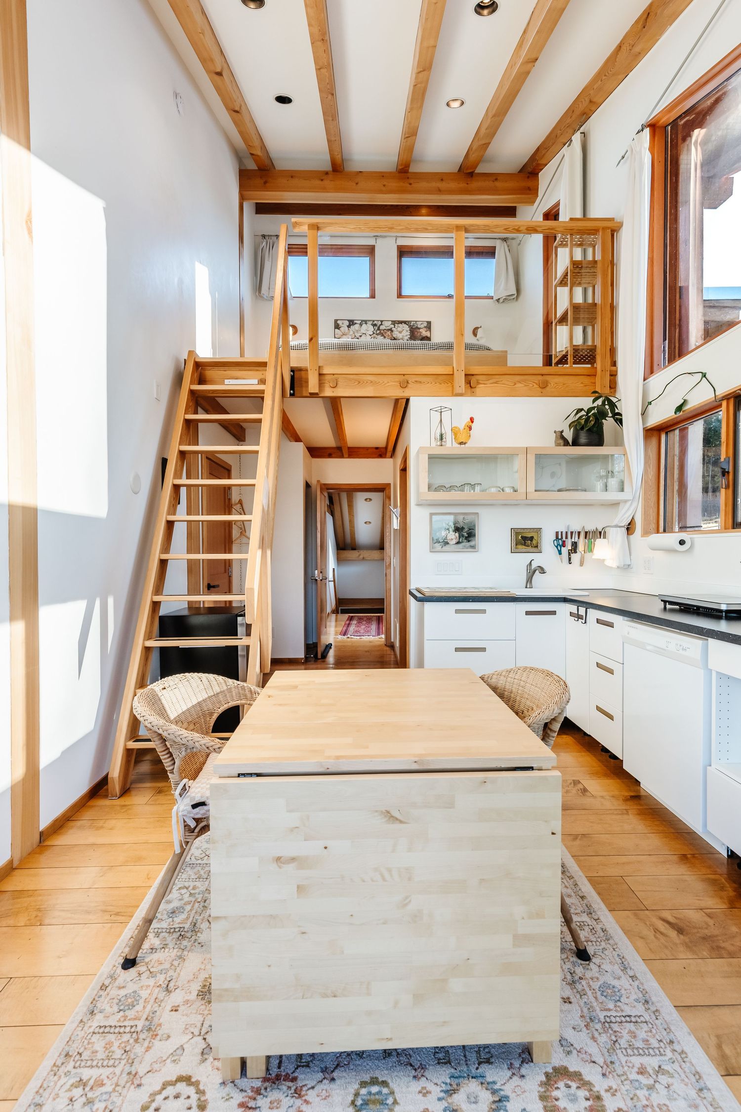 Modern tiny house interior with wooden beams, loft bedroom, white kitchen cabinets and rustic island on patterned rug.