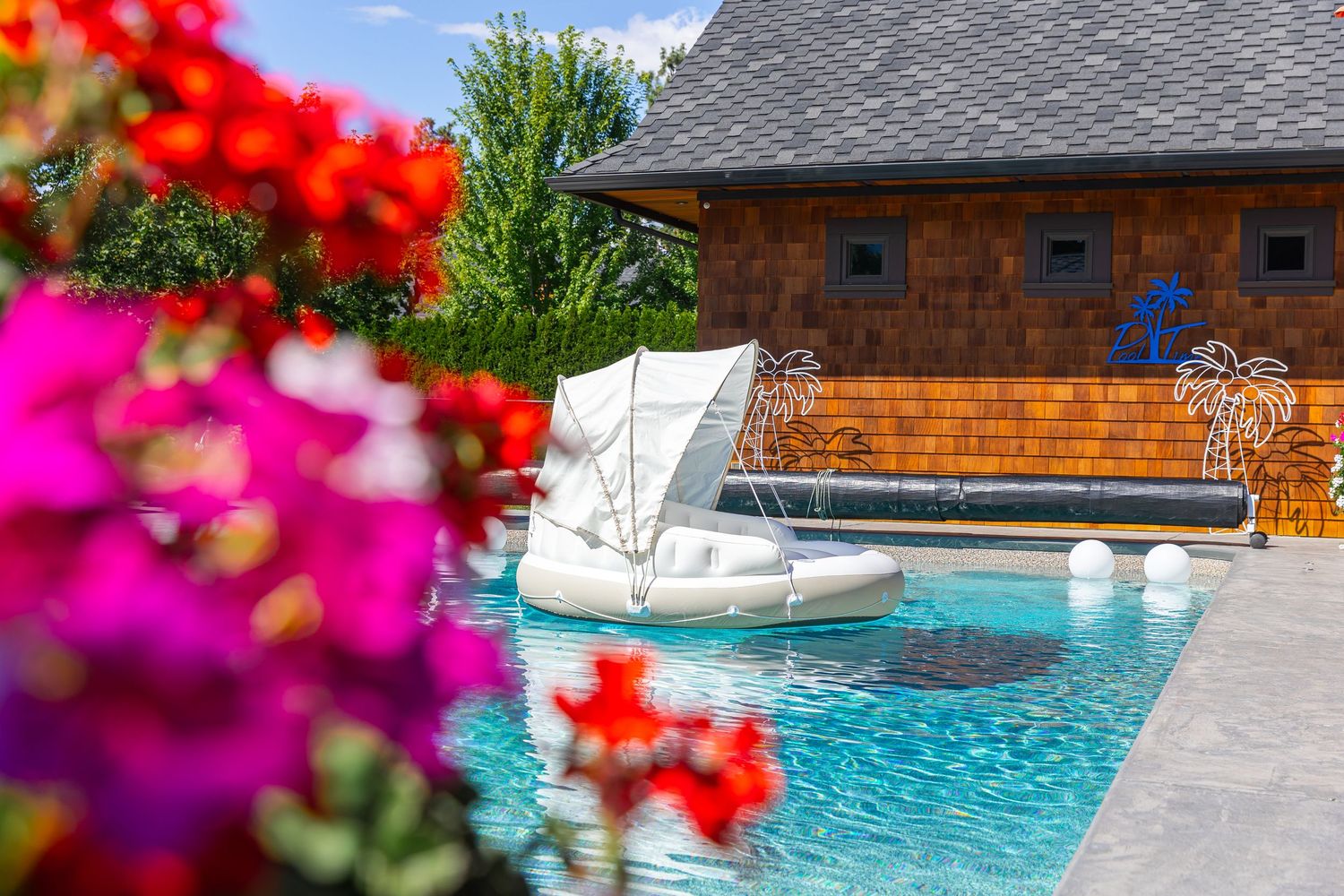 Vibrant red flowers frame a view of a backyard pool with swan floats and brick house.