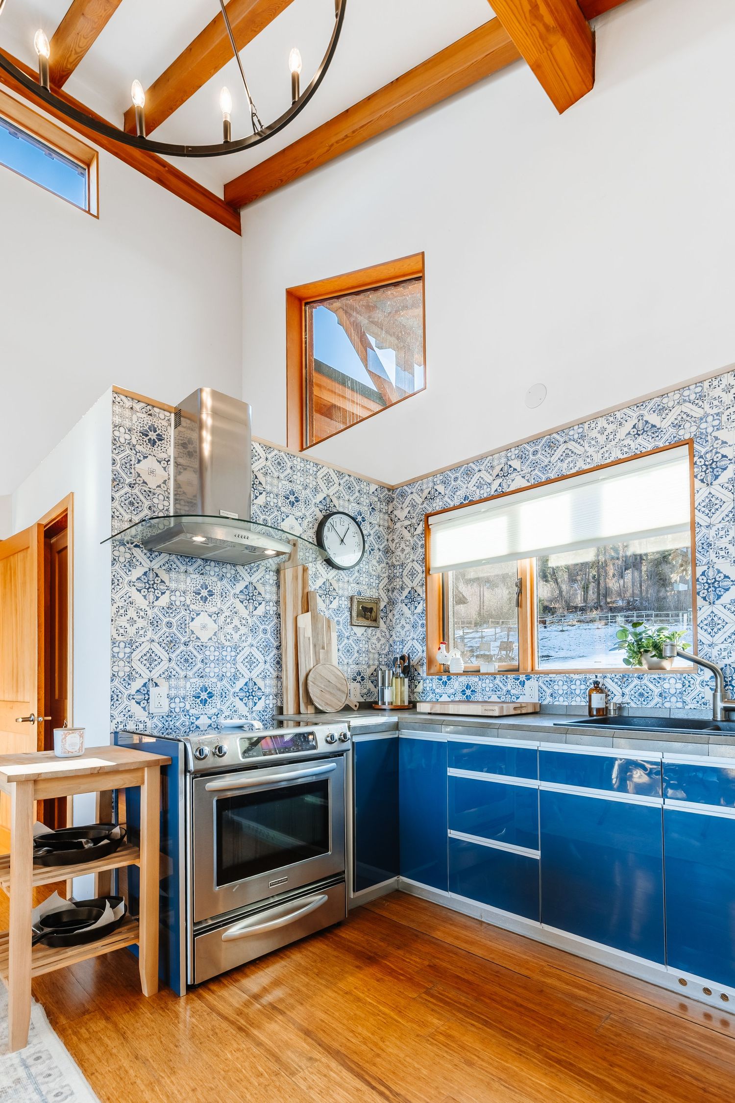 Modern kitchen with blue cabinets, wood beams, patterned wallpaper, and hardwood floors in an open-concept layout.