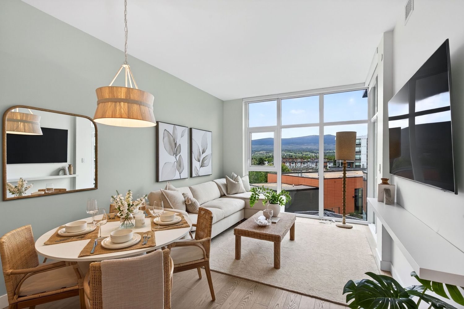 Modern apartment living room with sage green walls, large windows, mountain views, neutral sofa, and dining area with rattan chairs.