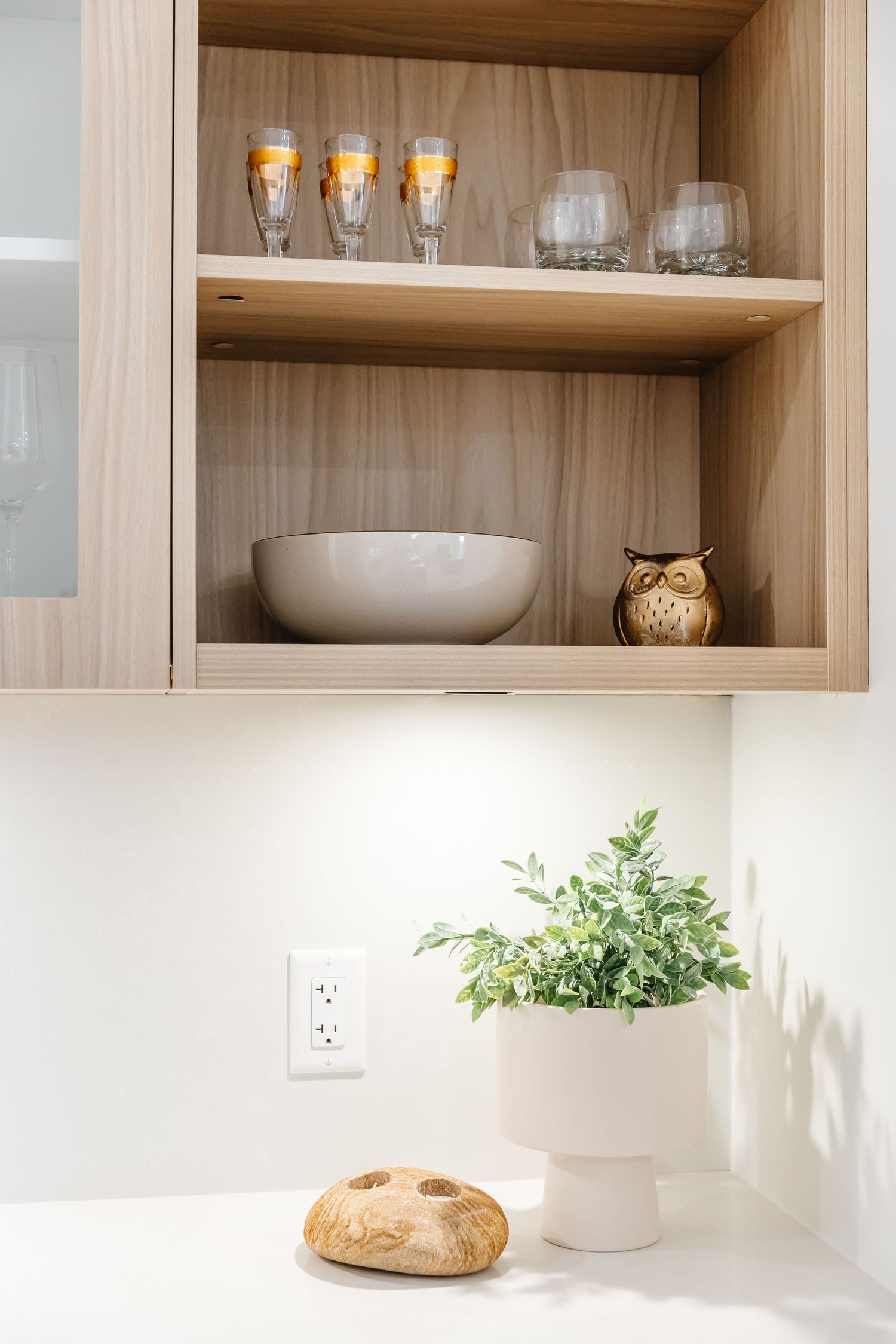 Modern kitchen shelving displays glassware and bowls with a potted plant and round bread loaf on white counter below.