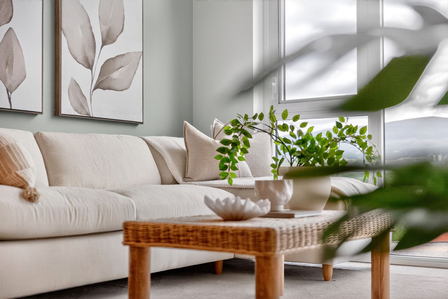 Cozy modern living room with white sofa, rattan coffee table, green plants, and botanical wall art in neutral tones.