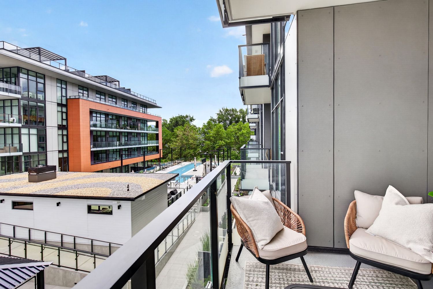 Modern apartment balcony with wicker chair and city views overlooking a pool and contemporary buildings on a sunny day.