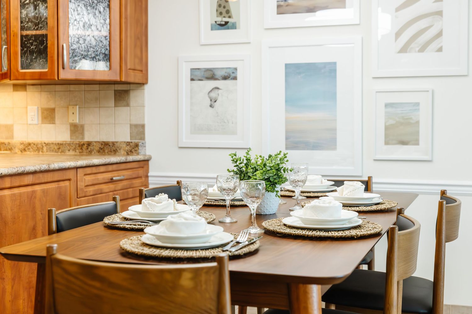 A bright dining room features white walls adorned with framed artwork and wooden cabinets along the perimeter.
