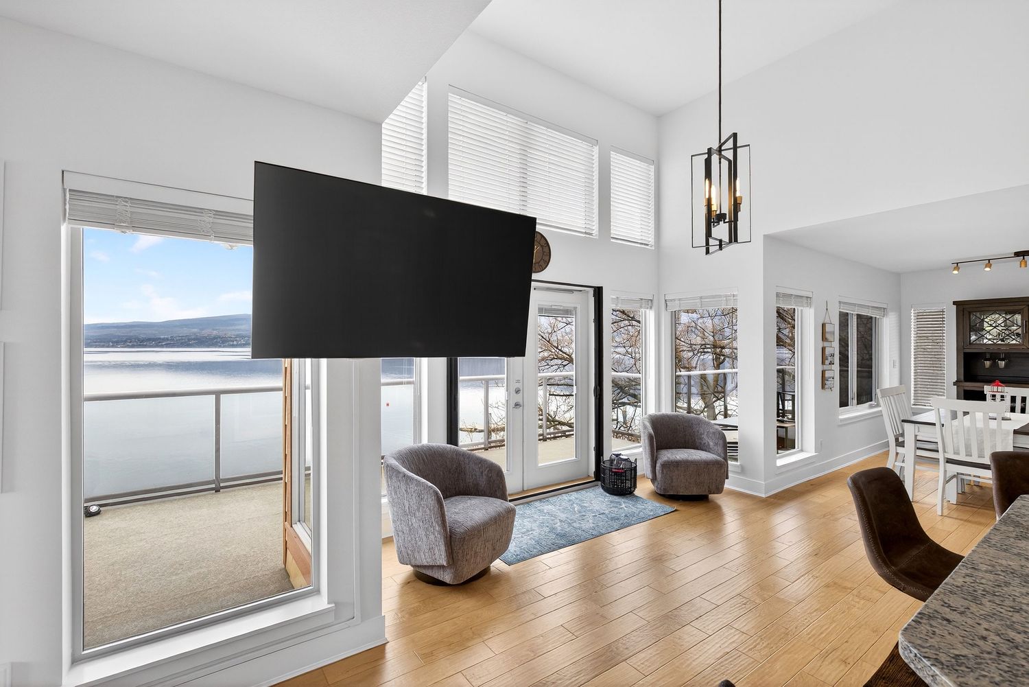 Bright living room with hardwood floors, wall-mounted TV, gray chairs, and panoramic water views through large windows.