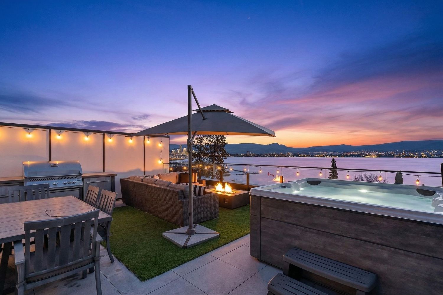 Rooftop patio with hot tub, fire pit, and outdoor seating overlooking a scenic waterfront at sunset.