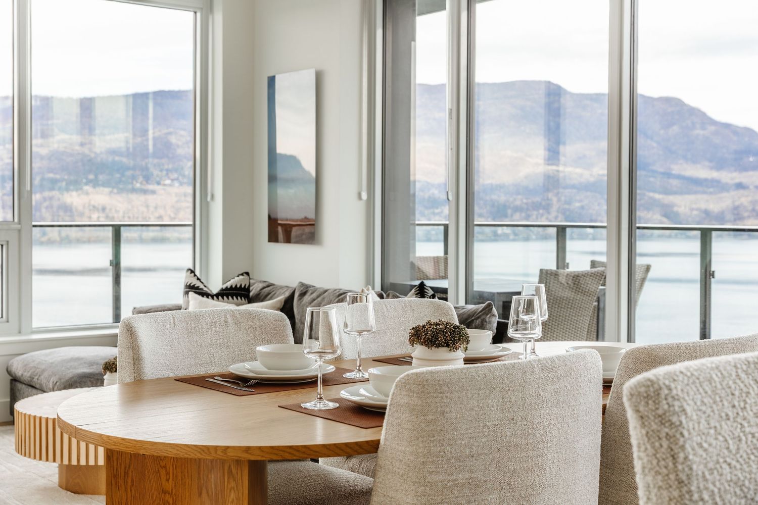 Modern dining table with cream chairs overlooking a scenic mountain lake view through large windows.