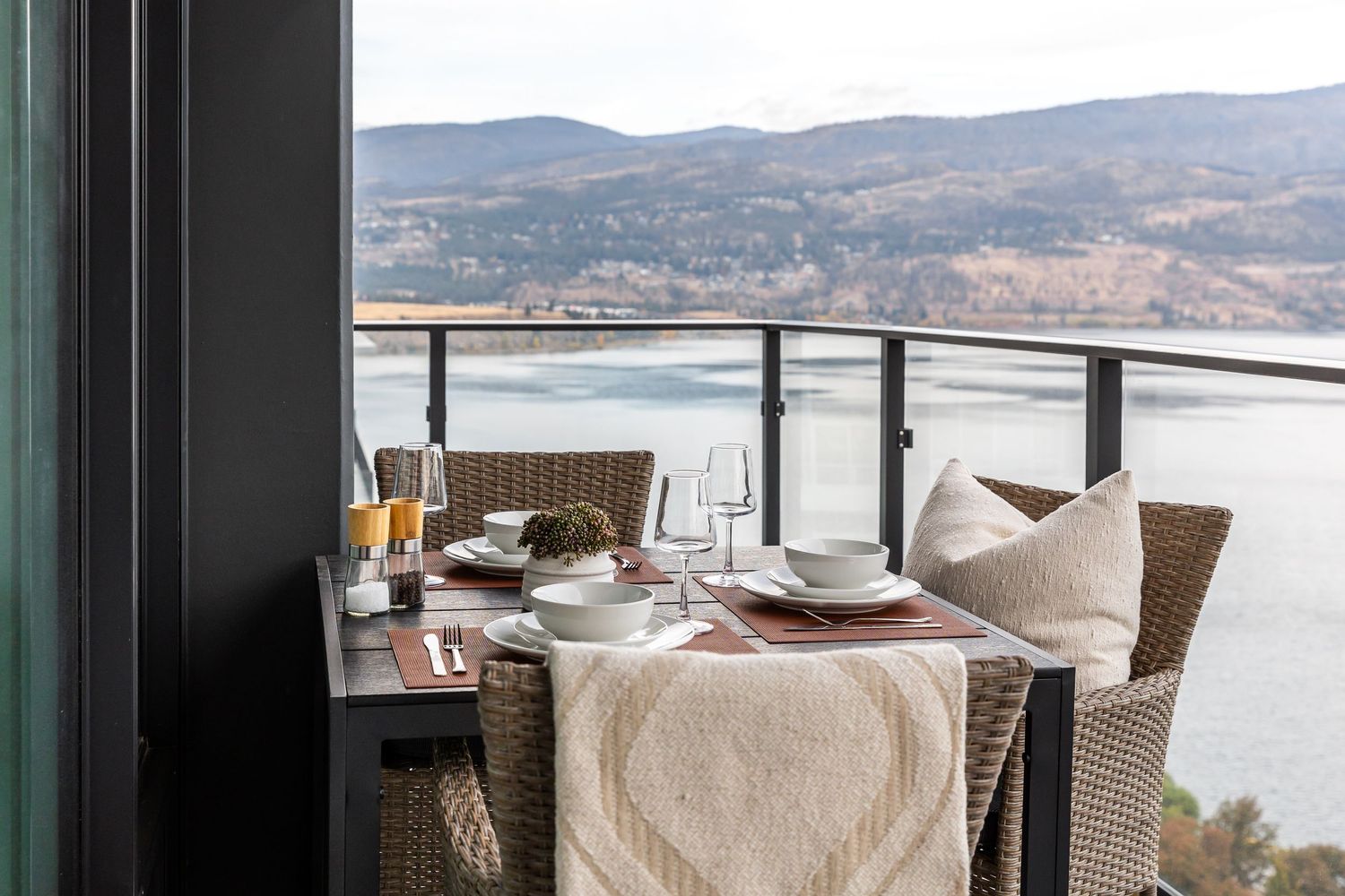 Outdoor dining table with wicker chairs on a balcony overlooking a scenic lake and mountain landscape.