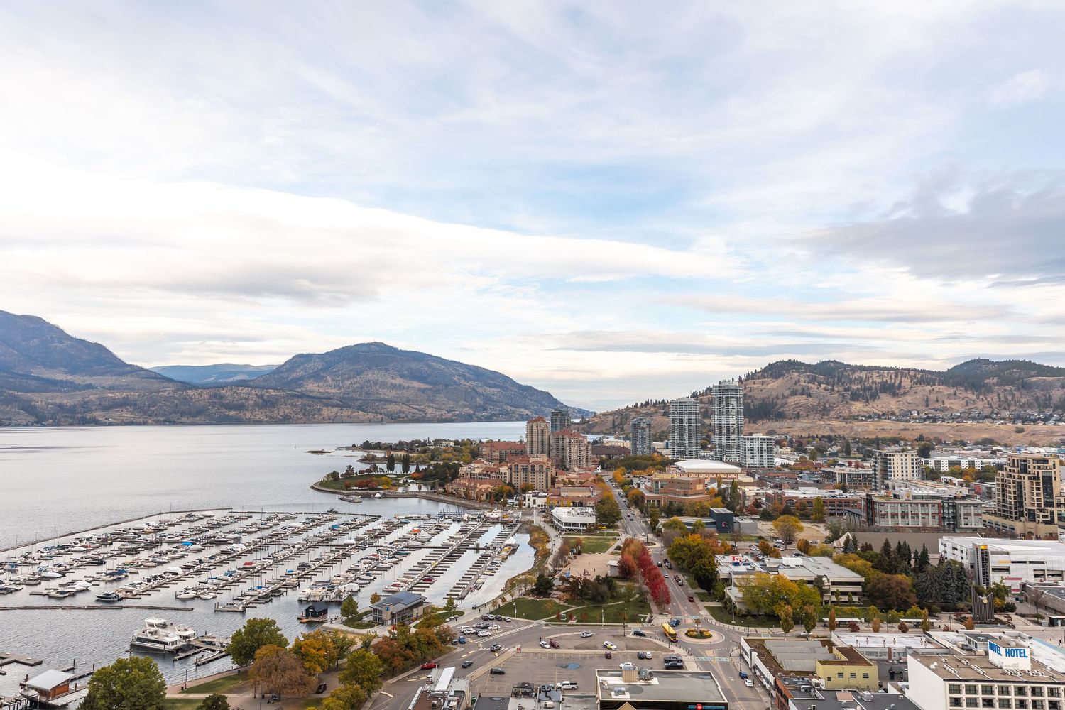 Panoramic aerial view of a coastal city with mountains, marina, and urban development along the Mediterranean shoreline.