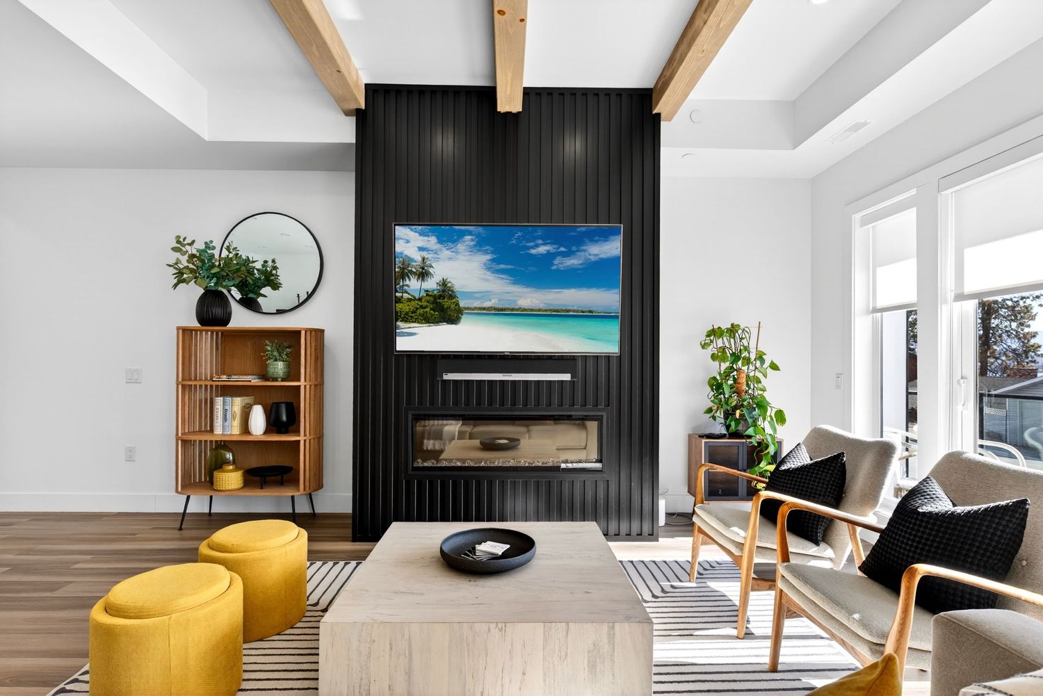 Modern living room with black slat fireplace wall, mounted TV, yellow ottomans, and wood beam ceiling.