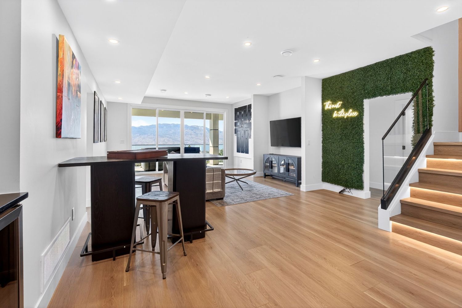 Modern open-plan living room with hardwood floors, green moss wall, neon sign, staircase, and bar seating area.