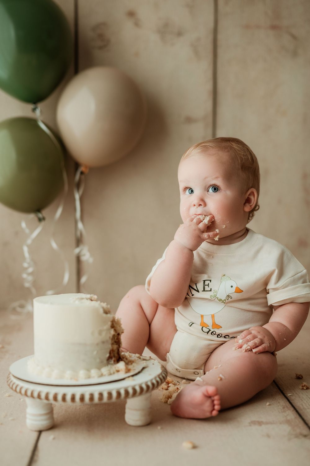 Baby in white outfit sitting next to a white birthday cake with green balloons in background.