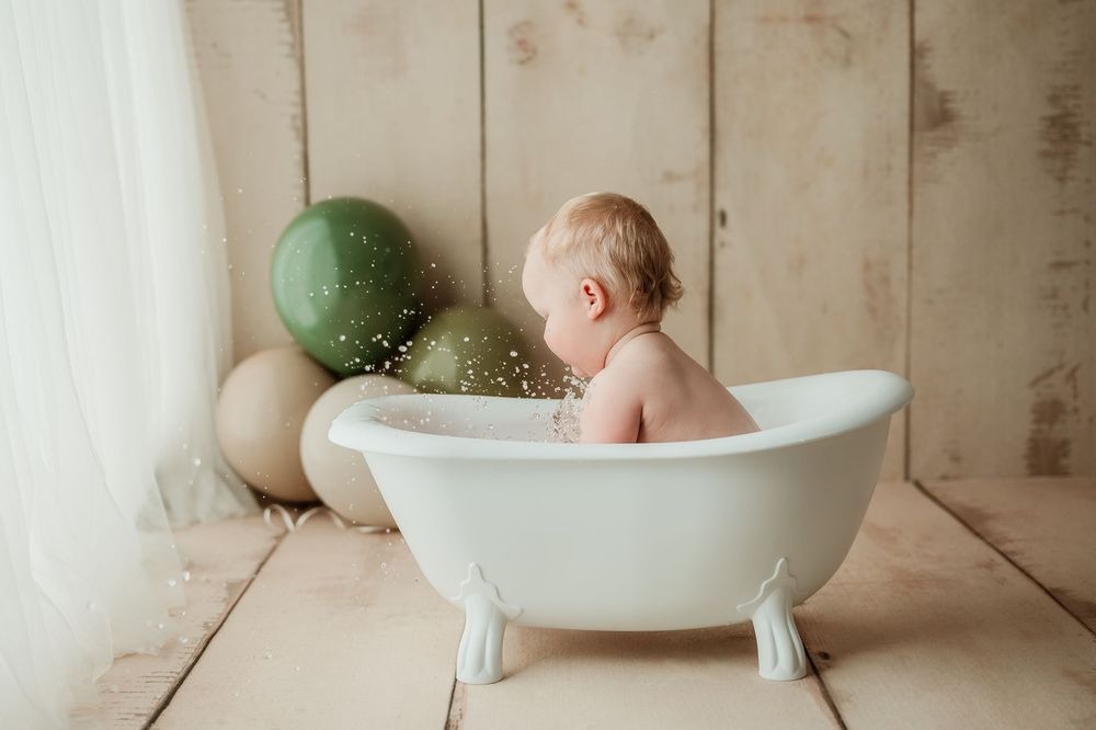 Baby bath photography session with white tub and green balloons on wooden backdrop.