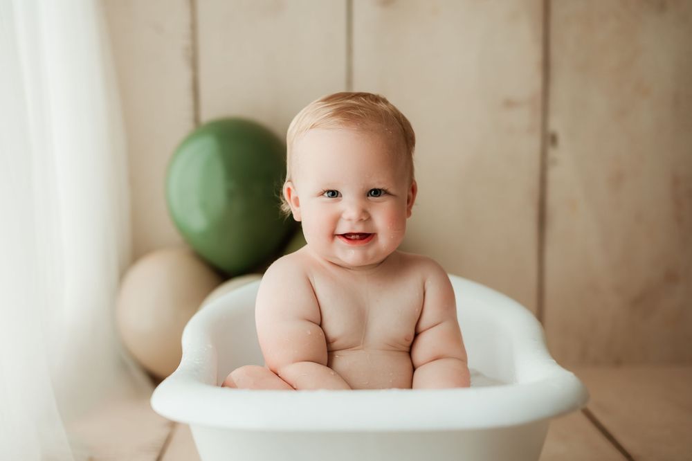 Happy baby sitting in white basin with green and neutral balloons in background.