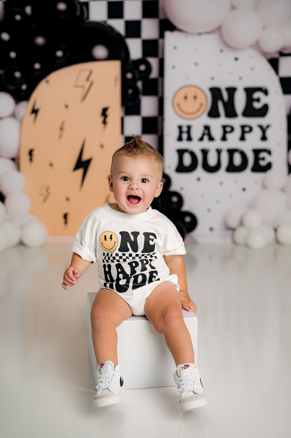 Baby wearing white ONE shirt and black shorts sits against white balloon backdrop with checkered pattern.