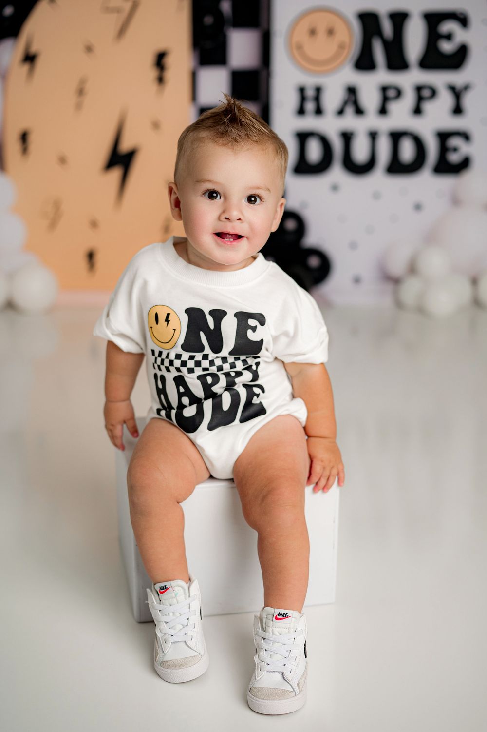 Baby wearing a 'Dude' graphic t-shirt and white sneakers sits on a grey background with decorative wall art.