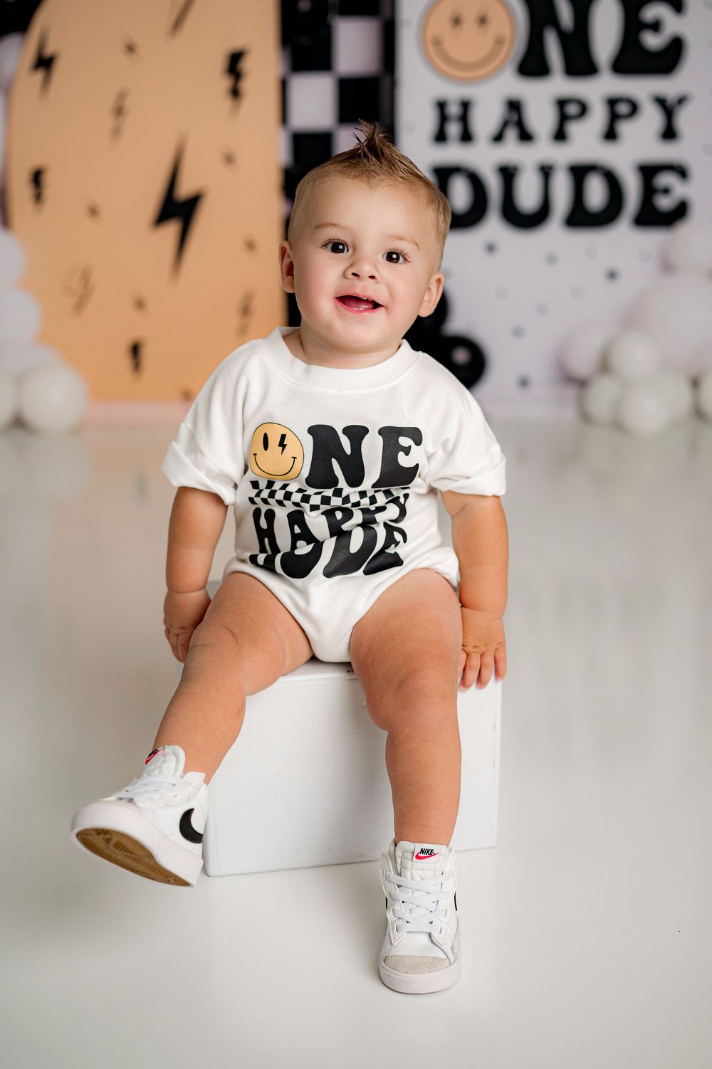 Baby in white onesie with smiley face design sits on white surface against backdrop with lightning bolt decorations.