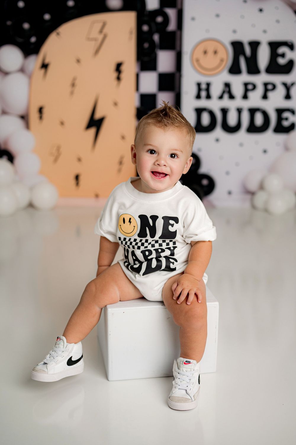 Adorable baby in white t-shirt and sneakers sits on white blocks against a festive party backdrop with lightning bolts and smileys.