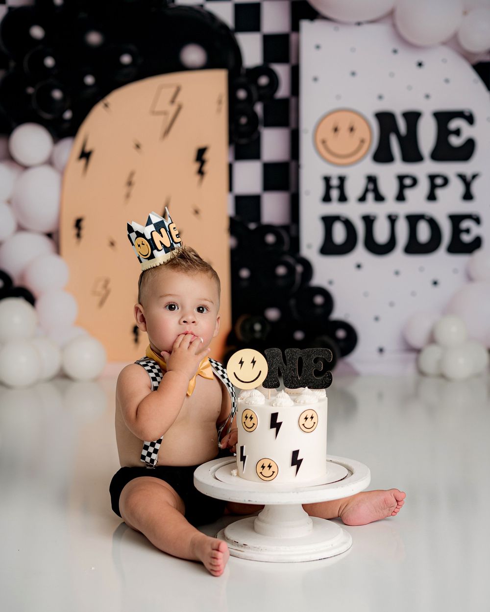 First birthday celebration setup with black and white decor, smiley faces, lightning bolts and a white cake on pedestal.