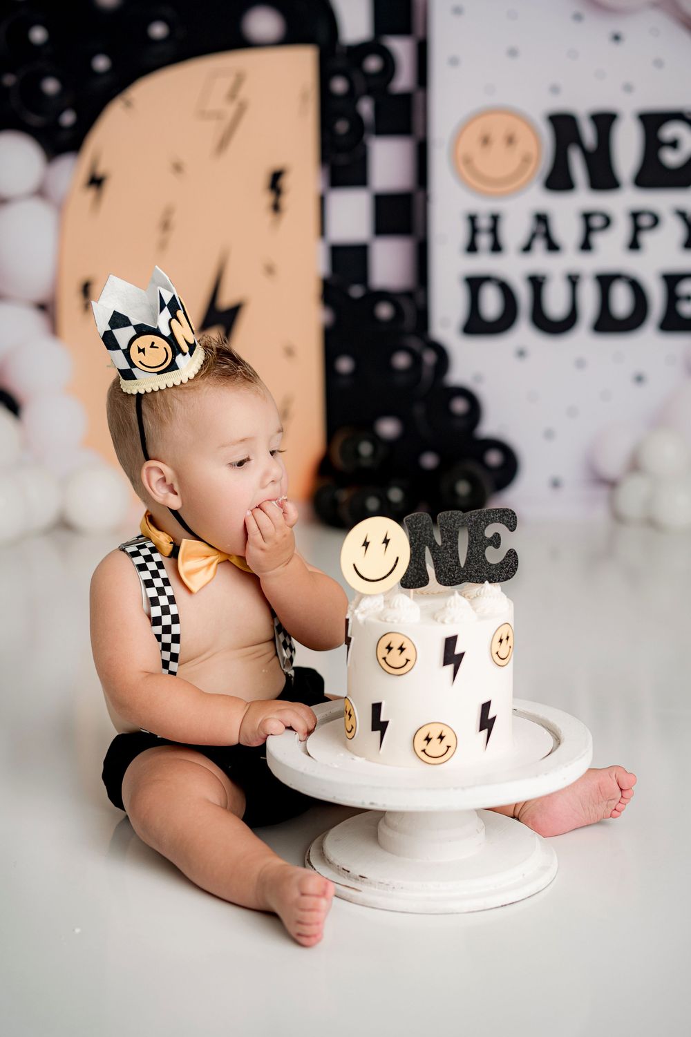 First birthday cake smash photo with black and white checkered theme and smiley face decorations on white cake.