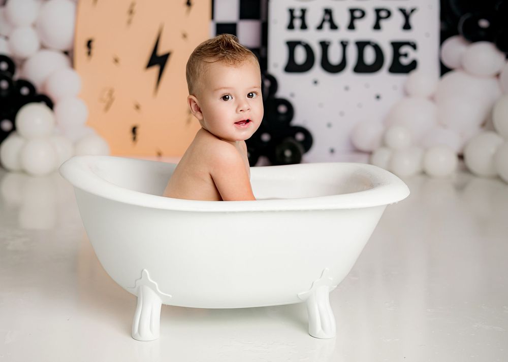 A small child sits in a white clawfoot bathtub against a festive black and white balloon backdrop with 'Happy Dude' text.