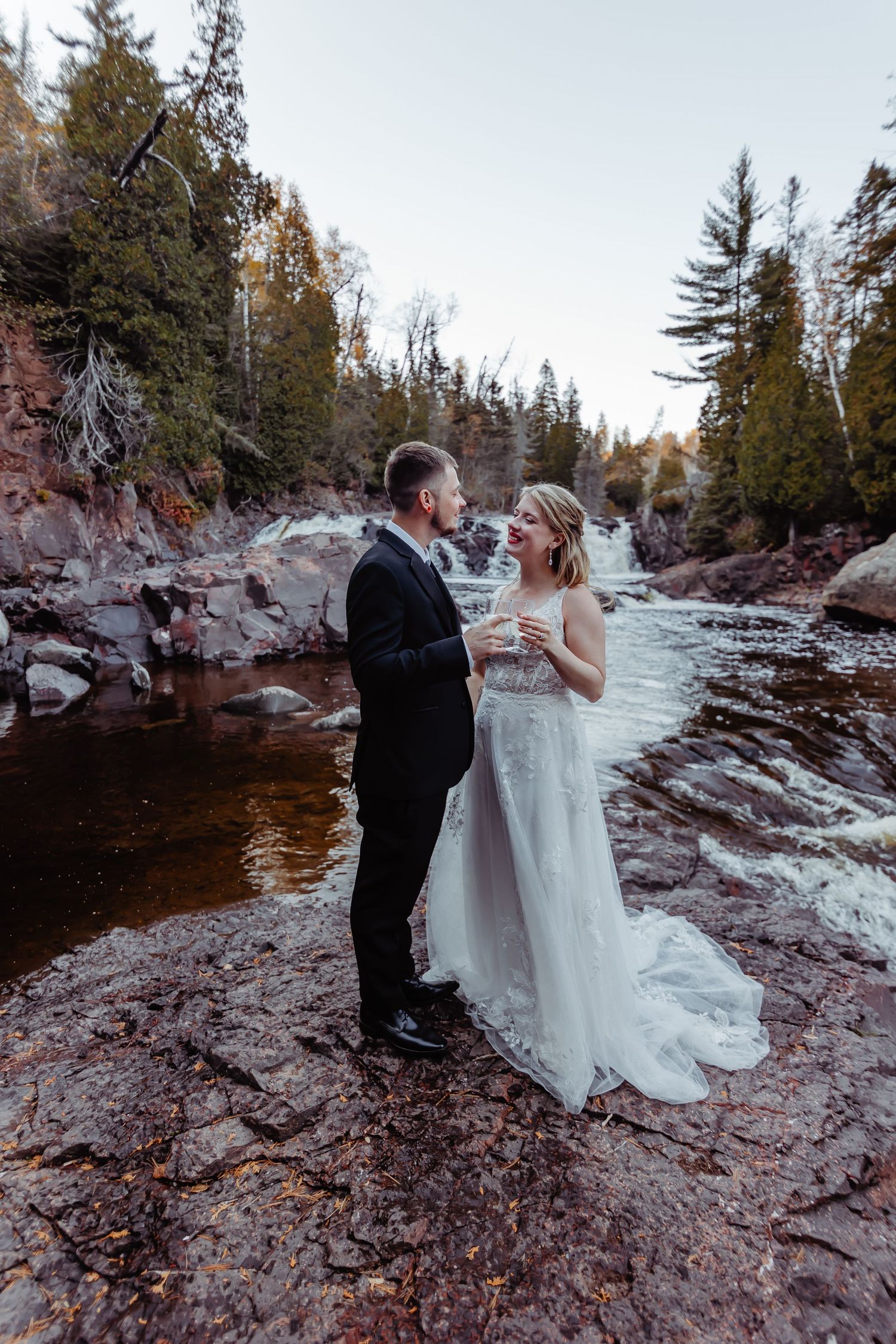 Couple in wedding attire standing on rocky riverbank with rushing water and evergreen trees.