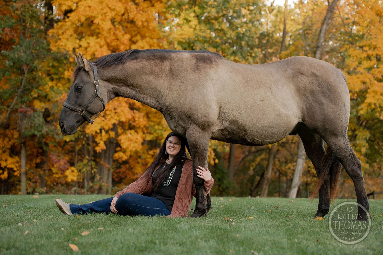 A gray horse walks past as someone sits on green grass with autumn colors in the background.